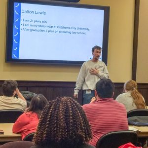 A young man in a Plan Your Way OK sweatshirt stands at the front of a classroom, pointing toward a large screen displaying a presentation.