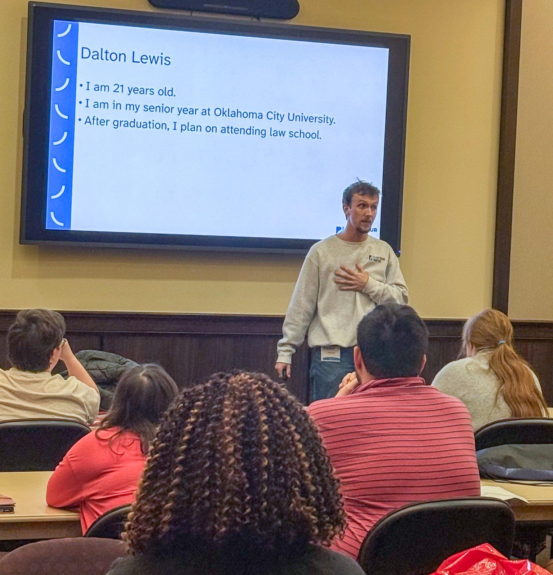 A young man in a Plan Your Way OK sweatshirt stands at the front of a classroom, pointing toward a large screen displaying a presentation.