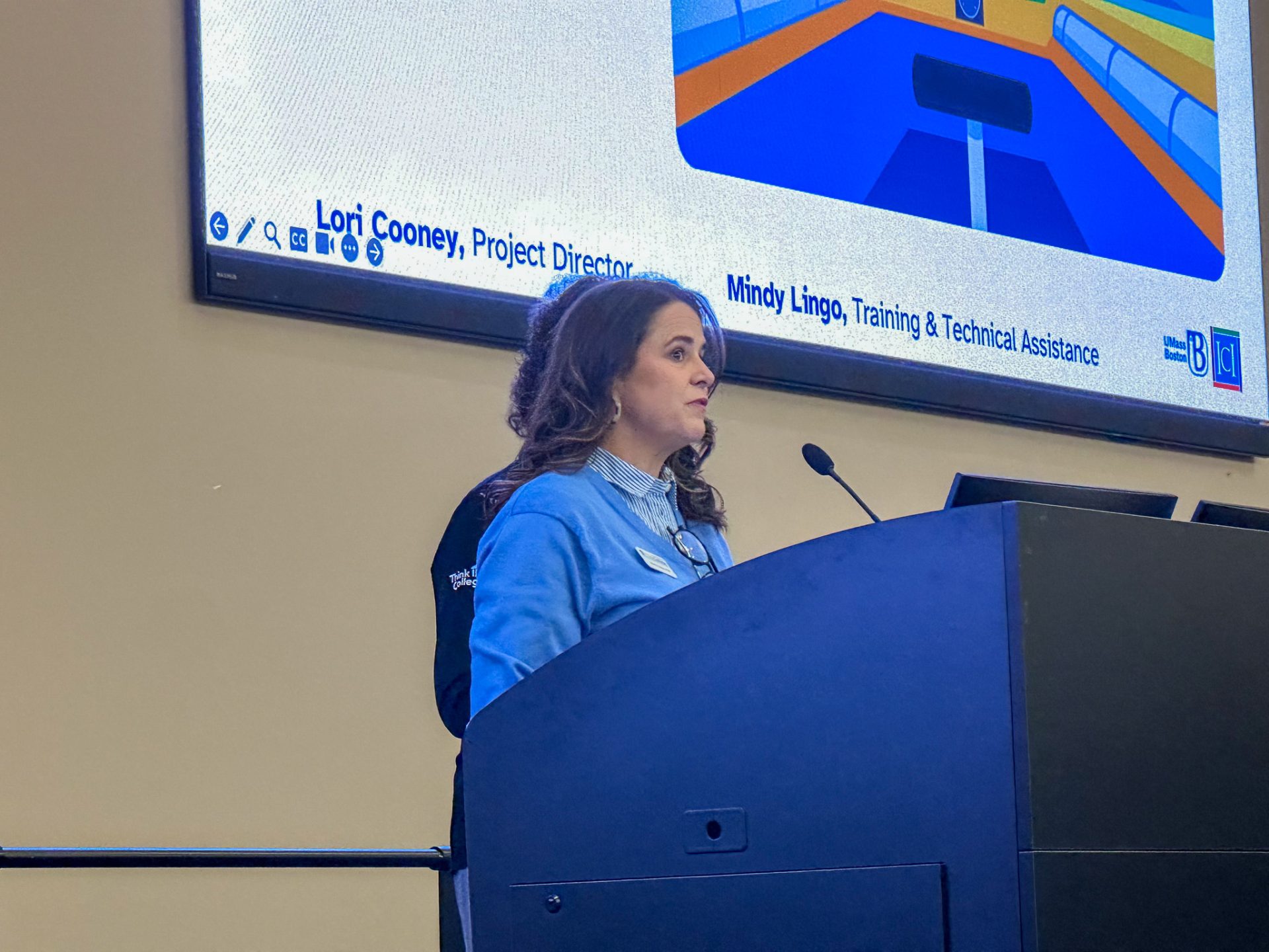 A woman in a light blue sweater stands behind a podium speaking with a large screen in the background.