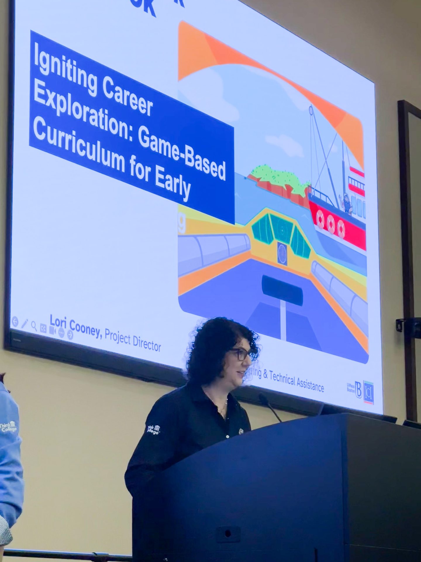 A woman with black curly hair stands at a podium in front of a large screen that reads "Igniting Career Exploration: Game Based Curriculum for Early"