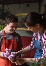 A boy with a red apron working with a female in a denim apron holding purple flowers