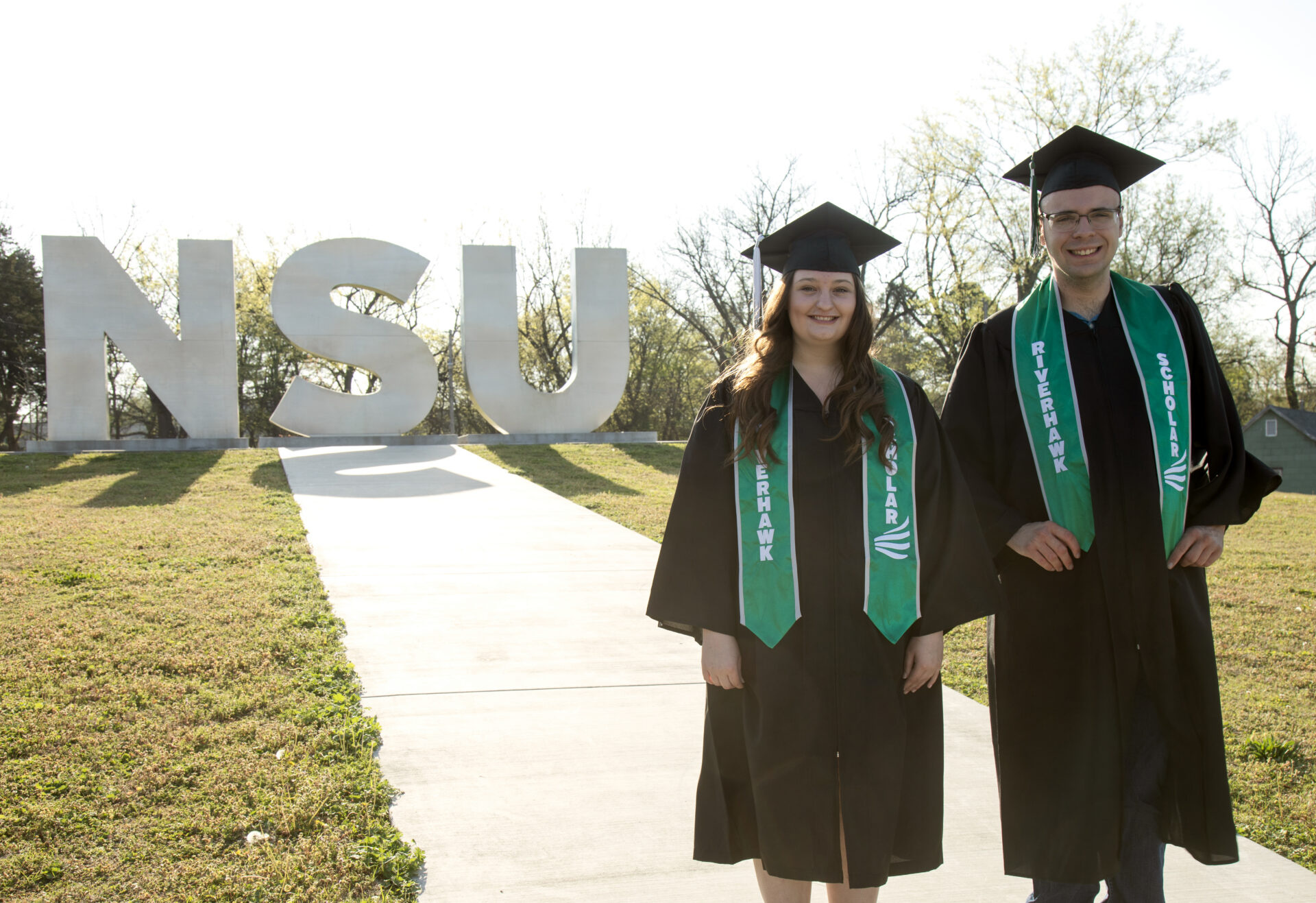 Riverhawk Scholars, Elizabeth and Micah standing on lawn wearing graduation cap and gown
